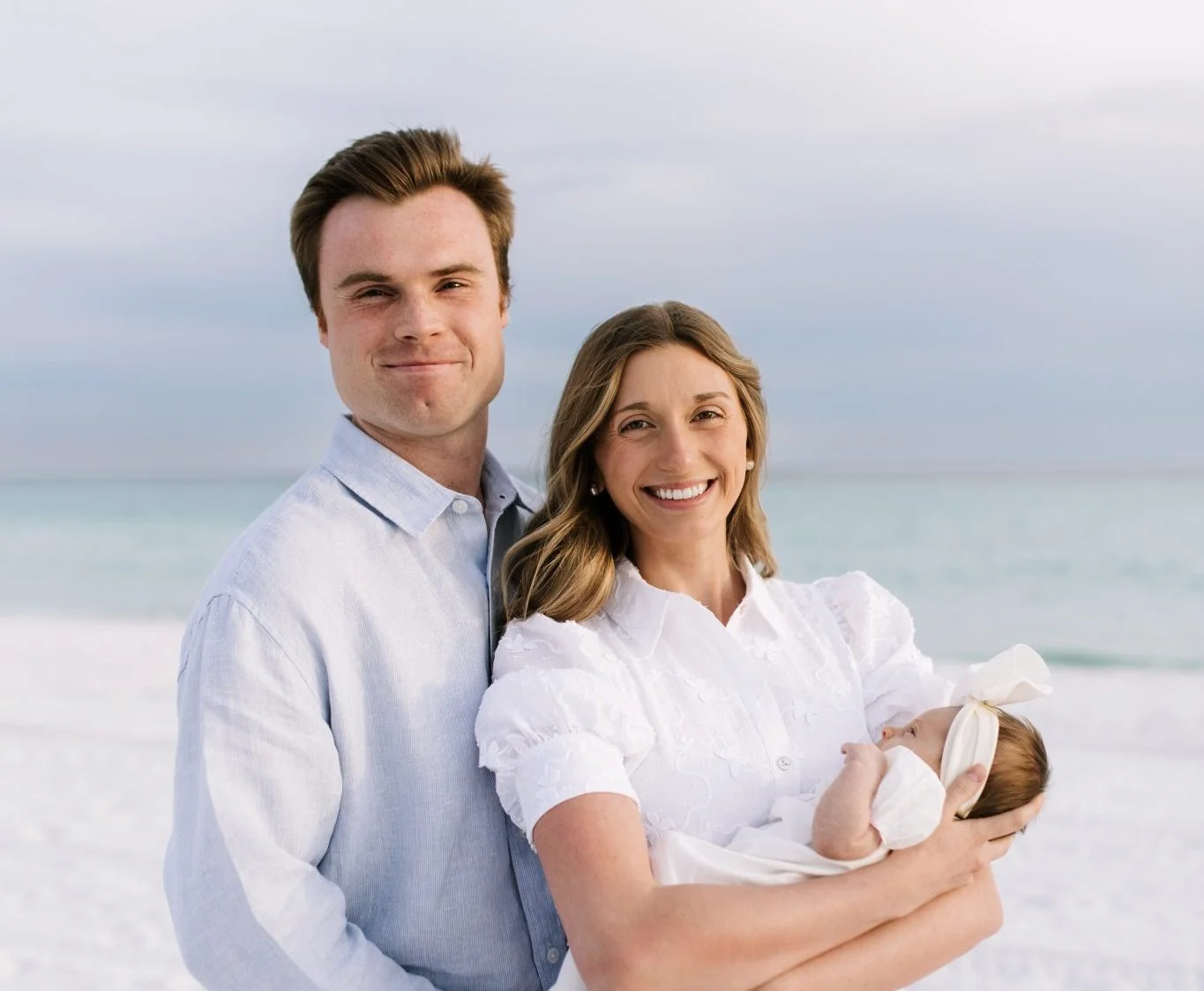 Smiling couple on a white-sand beach holding their newborn baby, with the sea in the background.